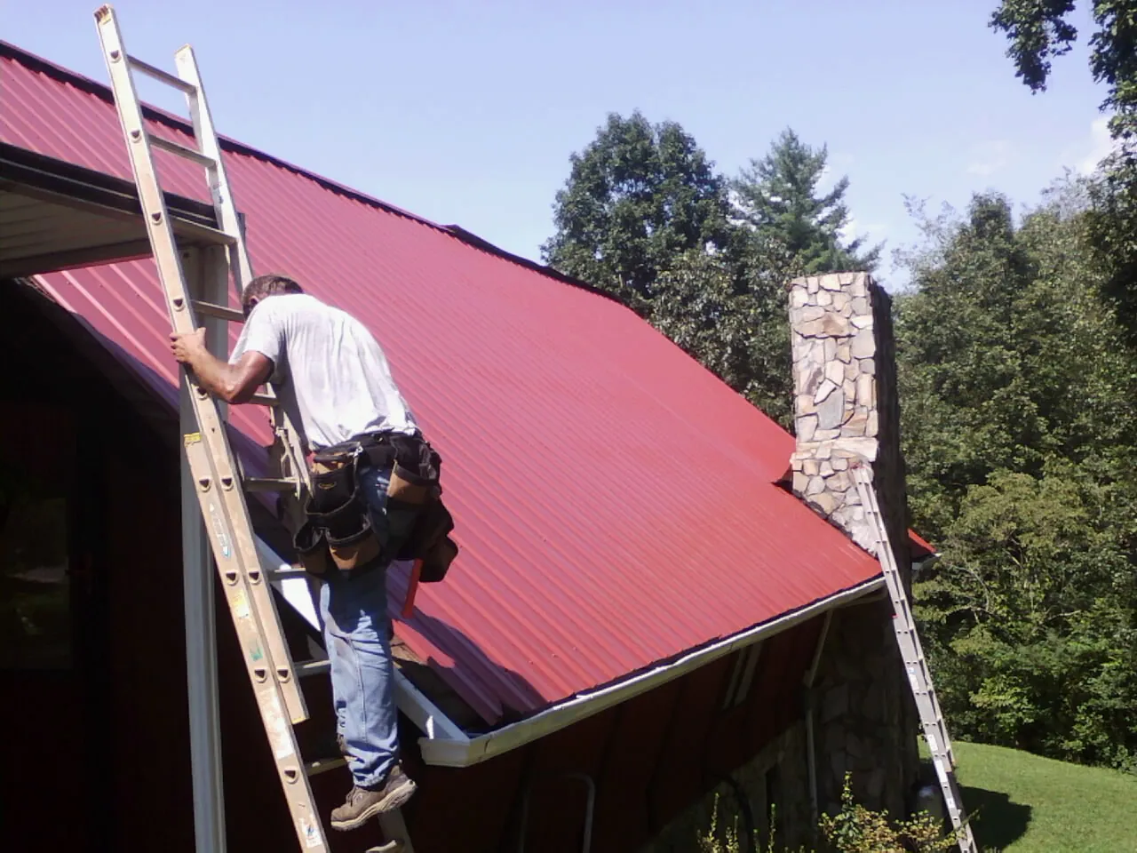 Roofer on a ladder beside a red metal roof and stone chimney — Soffit & Fascia Repair by Roofing Brothers in Fairfax